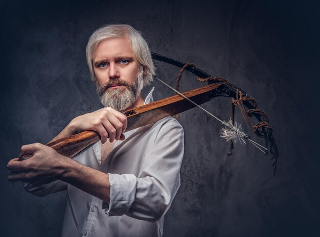 Handsome mature man with a gray beard and white shirt holding a crossbow on shoulder