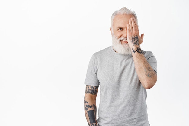 Handsome mature man with tattoos, hiding half of face, cover eye with hand and smiling happy at front, standing in grey t-shirt against white wall