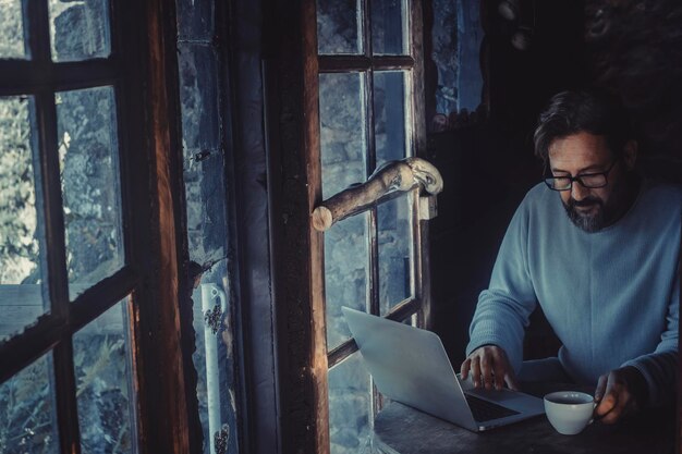 居心地​の​良い​木製​の​小屋​の​中​で​暗闇​の​中​で​ラップトップコンピューター​で​作業している​ハンサムな​成熟した​男性