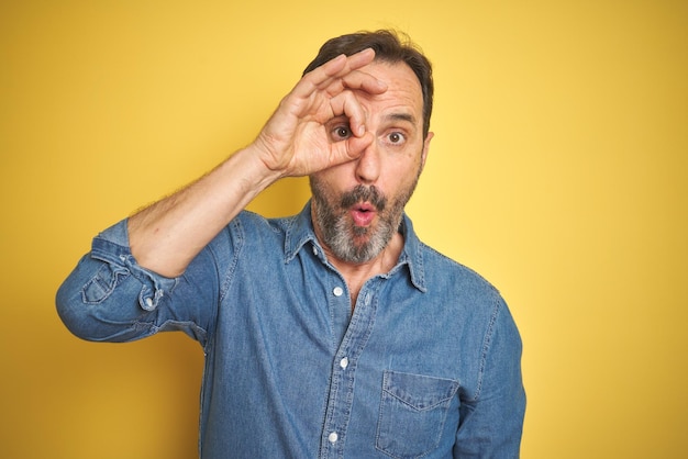 Foto gratuita bell'uomo anziano di mezza età con i capelli grigi su sfondo giallo isolato che fa un gesto ok scioccato con l'occhio del viso sorpreso che guarda attraverso le dita espressione incredula