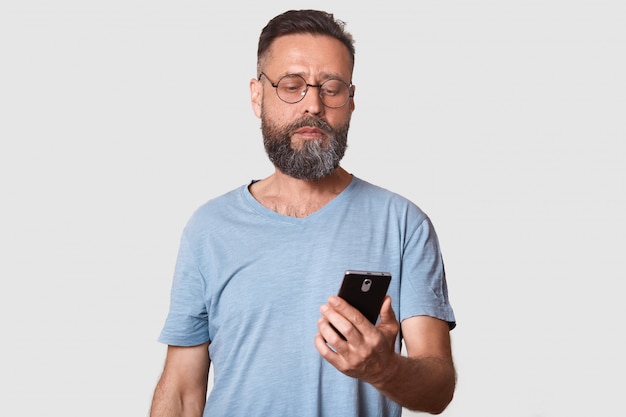 Handsome middle aged man using his phone with serious faxcial expression while standing against gray wall attractive male reading important message from wife. People and technology concept.