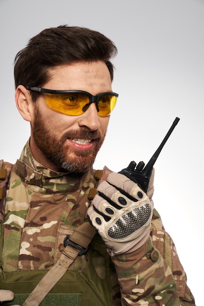 Бесплатное фото Красивый военный в желтых тактических очках разговаривает по рации, портрет улыбающегося солдата