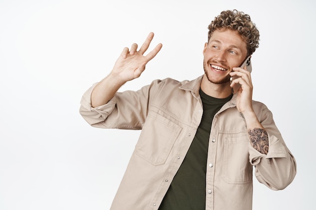 Handsome modern blond guy talking on mobile phone and saying hello to passerby Young man having a call and greeting someone with hand wave white background