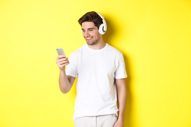 Handsome modern guy choosing playlist on smartphone, wearing headphones, standing over yellow background