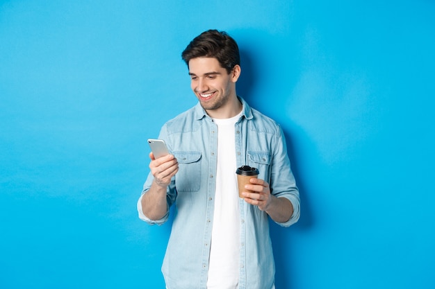 Красивый современный парень читает сообщение по мобильному телефону и пьет кофе, стоя на синем фоне