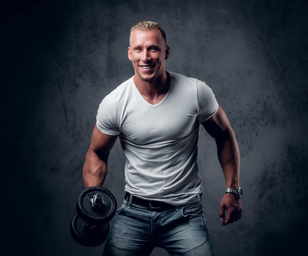 Handsome muscular blond male in a white t shirt holds dumbbell over grey artistic background.