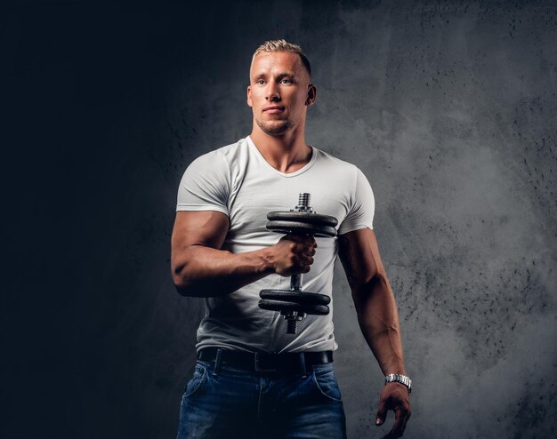 Handsome muscular blond male in a white t shirt holds dumbbell over grey artistic background.