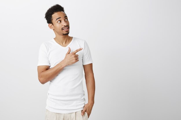 Handsome pleased dark-skinned guy in white t-shirt pointing upper right corner, making choice