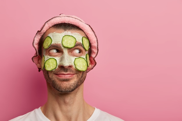 Handsome pleased man gets facial treatment, applies clay mask with slices of cucumbers, enjoys beauty procedures, has bristle