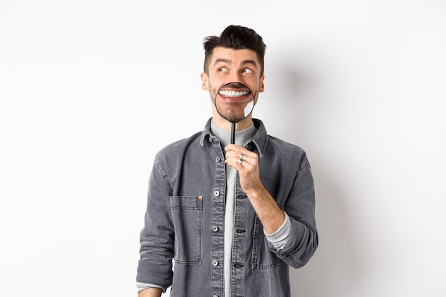 Handsome positive guy showing white perfect smile with magnifying glass, looking left at logo, standing against white background.