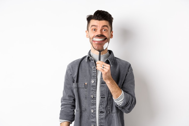 Handsome positive guy showing white perfect smile with magnifying glass, standing against white background
