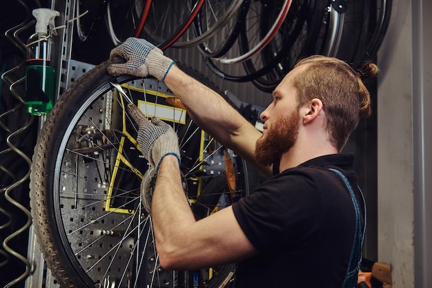 ジーンズのつなぎ服を着たハンサムな赤毛の男性。修理店で自転車のホイールを操作しています。作業員が作業場で自転車のタイヤを取り外します。