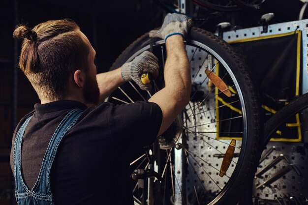 ジーンズのつなぎ服を着たハンサムな赤毛の男性。修理店で自転車のホイールを操作しています。作業員が作業場で自転車のタイヤを取り外します。