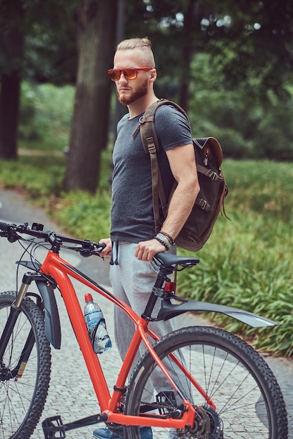 운동복과 선글라스를 쓴 세련된 머리와 수염을 가진 잘생긴 빨간 머리 남성은 자전거와 배낭을 메고 공원을 걷습니다.