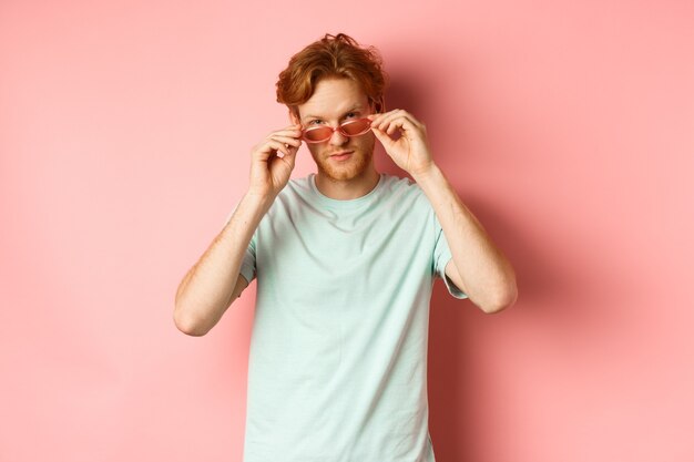 ピンクの背景の上に立っているカメラで生意気に見える夏のサングラスでハンサムな赤毛の男