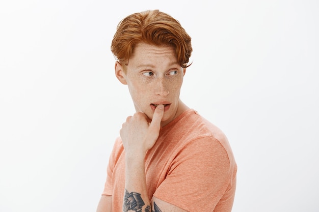 Handsome redhead man with tattoos looking intrigued to the right, thinking over white wall