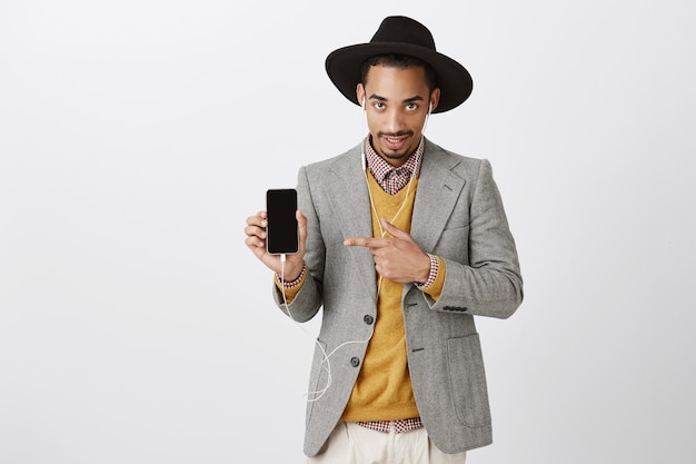 Handsome sassy Black guy in suit and hipster hat, listening music in earphones, pointing finger at smartphone screen