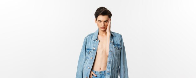 Красивый и нахальный гей в джинсовой куртке на голом туловище трогает лицо и выглядит уверенно