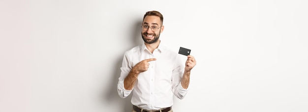 Бесплатное фото Красивый довольный мужчина в очках, указывающий на кредитную карту, довольный банковскими услугами, стоящими над w