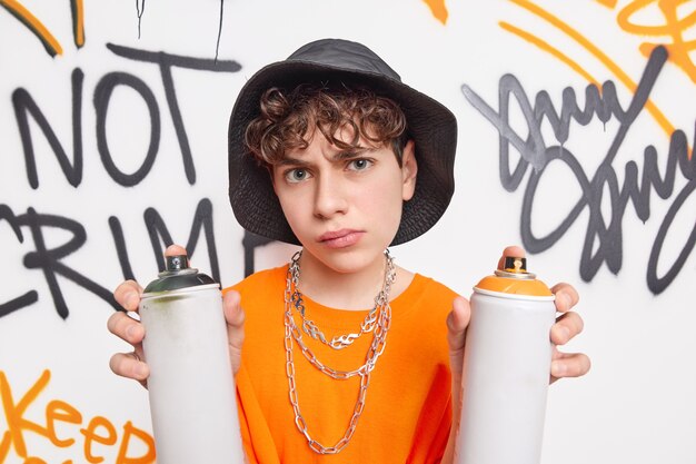 Красивый школьник очень серьезно смотрит в камеру, проводит свободное время после школы с друзьями, рисует граффити стену с помощью аэрозольных баллончиков, носит шляпу оранжевая футболка с металлическими цепями