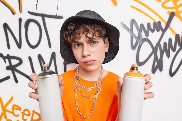 Handsome schoolboy looks very serious at camera spends free time after school with friends drawing graffiti wall with aerosol sprays wears hat orange t shirt metal chains