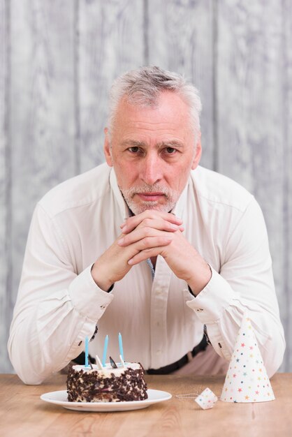 Handsome senior man looking at camera with delicious cake and party hat on table