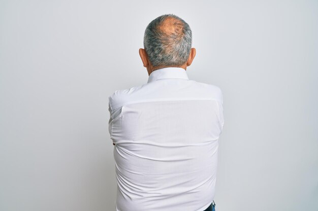 Красивый пожилой мужчина в повседневной белой рубашке стоит задом наперёд и смотрит в сторону со скрещенными руками