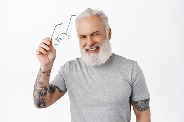 Handsome senior man with tattoos take off glasses laughing and smiling happy Stylish hipster old guy looking cheerful at camera standing over white background