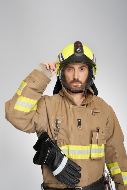Free photo handsome serious firefighter in uniform putting on protective helmet indoors portrait of bearded