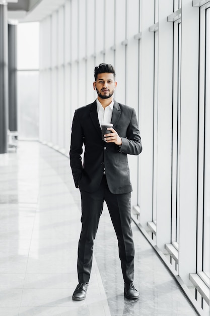 Handsome serious smiling Indian man in black suit in modern office center holding coffee in hands