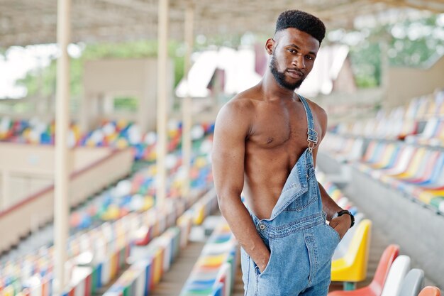 Красивый сексуальный афроамериканец с обнаженным туловищем в джинсовом комбинезоне позирует на цветных стульях на стадионе Модный портрет чернокожего мужчины