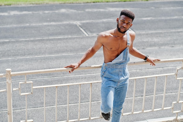 Handsome sexy african american bare torso man at jeans overalls posed at stadium Fashionable black man portrait