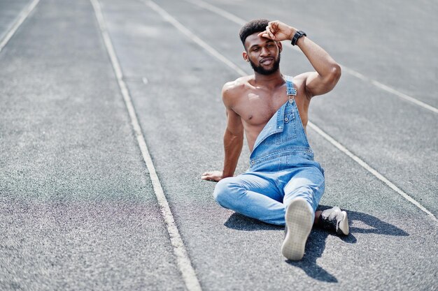 スタジアムの競馬場に座っているジーンズのオーバーオールでハンサムなセクシーなアフリカ系アメリカ人の裸の胴体の男ファッショナブルな黒人男性の肖像画