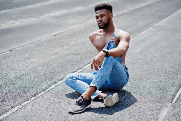Красивый сексуальный афроамериканец с голым туловищем в джинсовом комбинезоне сидит на ипподроме стадиона Модный портрет чернокожего мужчины