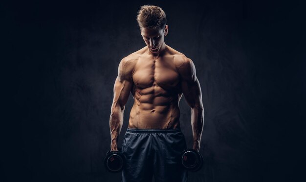 Handsome shirtless man with stylish hair and muscular ectomorph body doing the exercises with dumbbells on a dark textured background.
