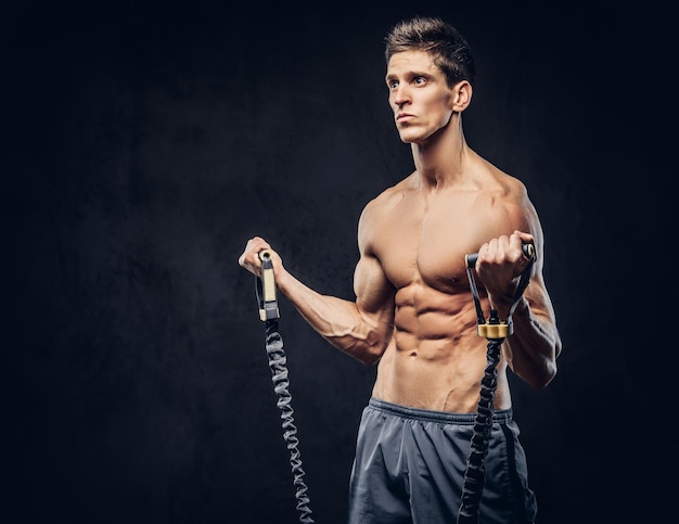 Handsome shirtless man with stylish hair and muscular ectomorph doing the exercises with an expander on a dark textured background.
