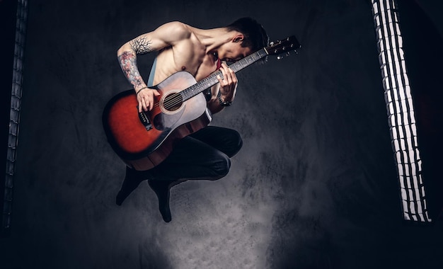 ジャンプしながらギターを弾くハンサムな上半身裸の若いミュージシャン。暗い背景に分離されています。