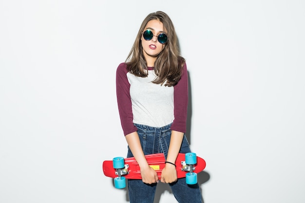 Handsome skater girl in casual clothes and black sunglasses holding red skateboard isolated on white wall