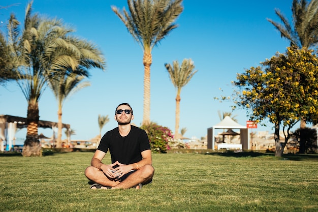 Free photo handsome slim bearded man in sunglasses sitting on green grass and relax enjoy summer vacation.