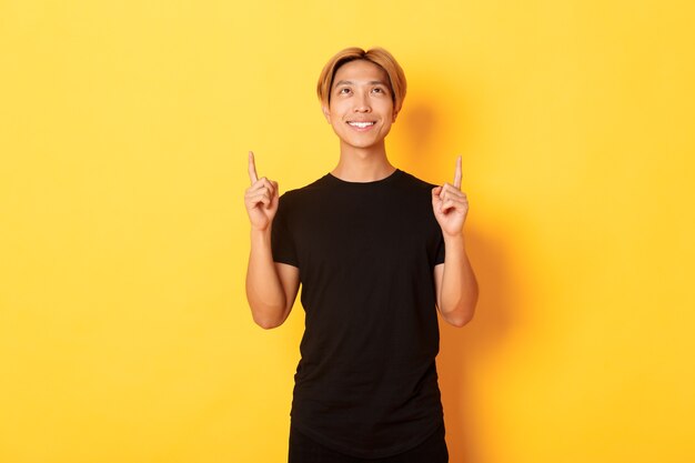 Handsome smiling asian man in black t-shirt pointing fingers up, yellow wall.