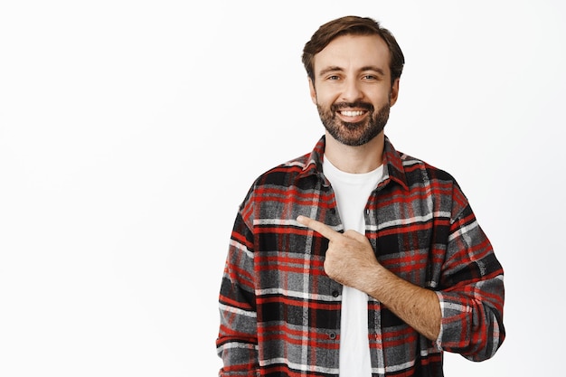 Handsome smiling bearded guy 30 years old pointing finger left showing advertisement standing over white background