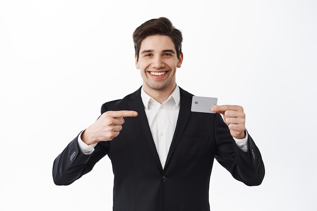 Красивый улыбающийся предприниматель указывает на кредитную карту, рекомендует банк, продвигает новую функцию, стоит на белом фоне в строгом костюме
