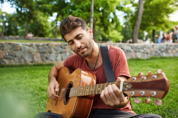 평온한 주말을 보내고 잔디에 앉아 공원에서 기타를 연주 잘 생긴 웃는 남자