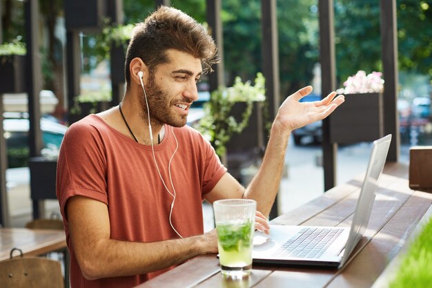 Handsome smiling guy talking with coworkers, using laptop video call application and earphones while sitting outdoor cafe