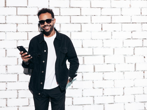 Bello sorridente modello hipster uomo sexy con la barba lunga vestito in camicia estiva nera e jeans vestiti moda maschio in posa vicino al muro di mattoni bianchi in studio al giorno di sole isolato