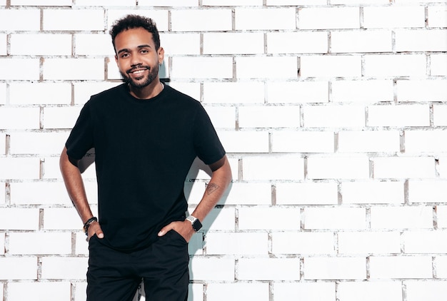 Handsome smiling hipster model Sexy unshaven man dressed in black summer tshirt and jeans clothes Fashion male posing near white brick wall in studio at sunny day Isolated