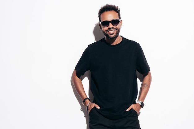 Handsome smiling hipster model Sexy unshaven man dressed in black summer tshirt and jeans clothes Fashion male posing near white wall in studio at sunny day Isolated