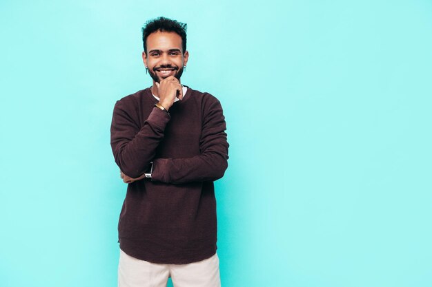 Handsome smiling hipster model Sexy unshaven man dressed in sweater and jeans clothes Fashion male posing near blue wall in studioLooking at camera and touching his chin