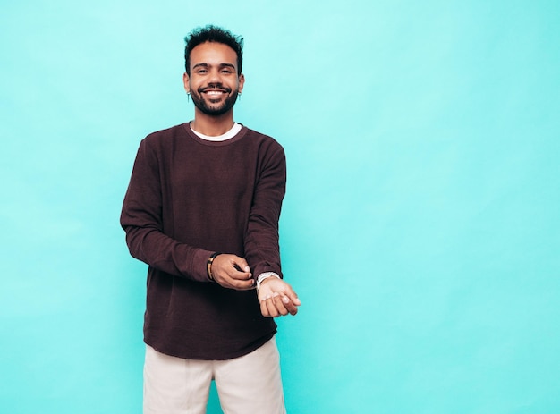 Handsome smiling hipster model Sexy unshaven man dressed in sweater and jeans clothes Fashion male posing near blue wall in studioLooking at camera