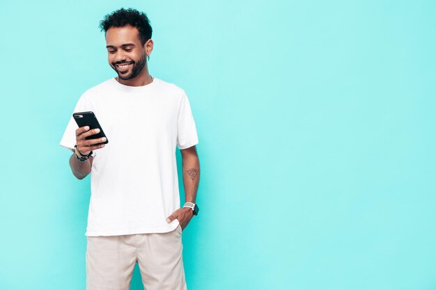 Handsome smiling hipster model Sexy unshaven man dressed in white summer tshirt and jeans clothes Fashion male posing near blue wall Holding smartphone Looking at cellphone screen Using apps